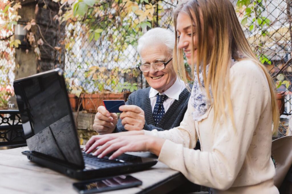 A grandparent and young adult grandchild sitting together with a laptop or tablet, smiling and interacting naturally.
